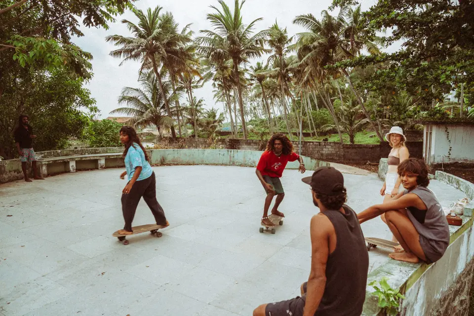 Skaters riding a concrete bowl surrounded by palm trees while friends watch from the edge