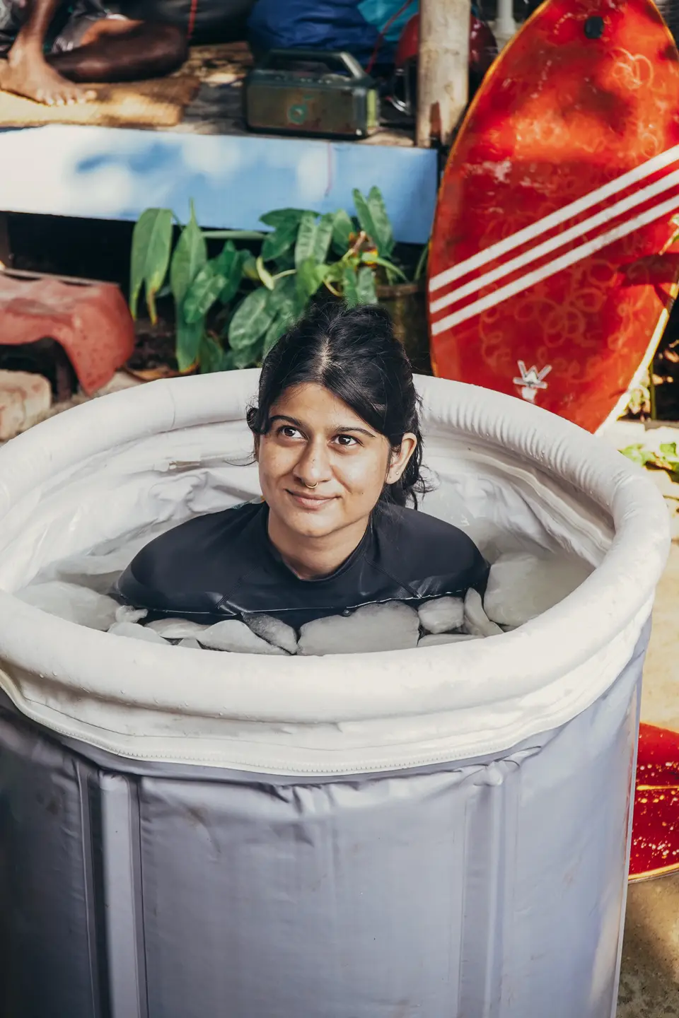 A smiling woman sitting in an ice bath full of ice, surfboards and plants behind her